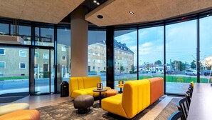 A modern hotel lobby with large windows, yellow sofas, a wooden reception desk, and a patterned carpet. - harry's home Salzburg (Salzburg)