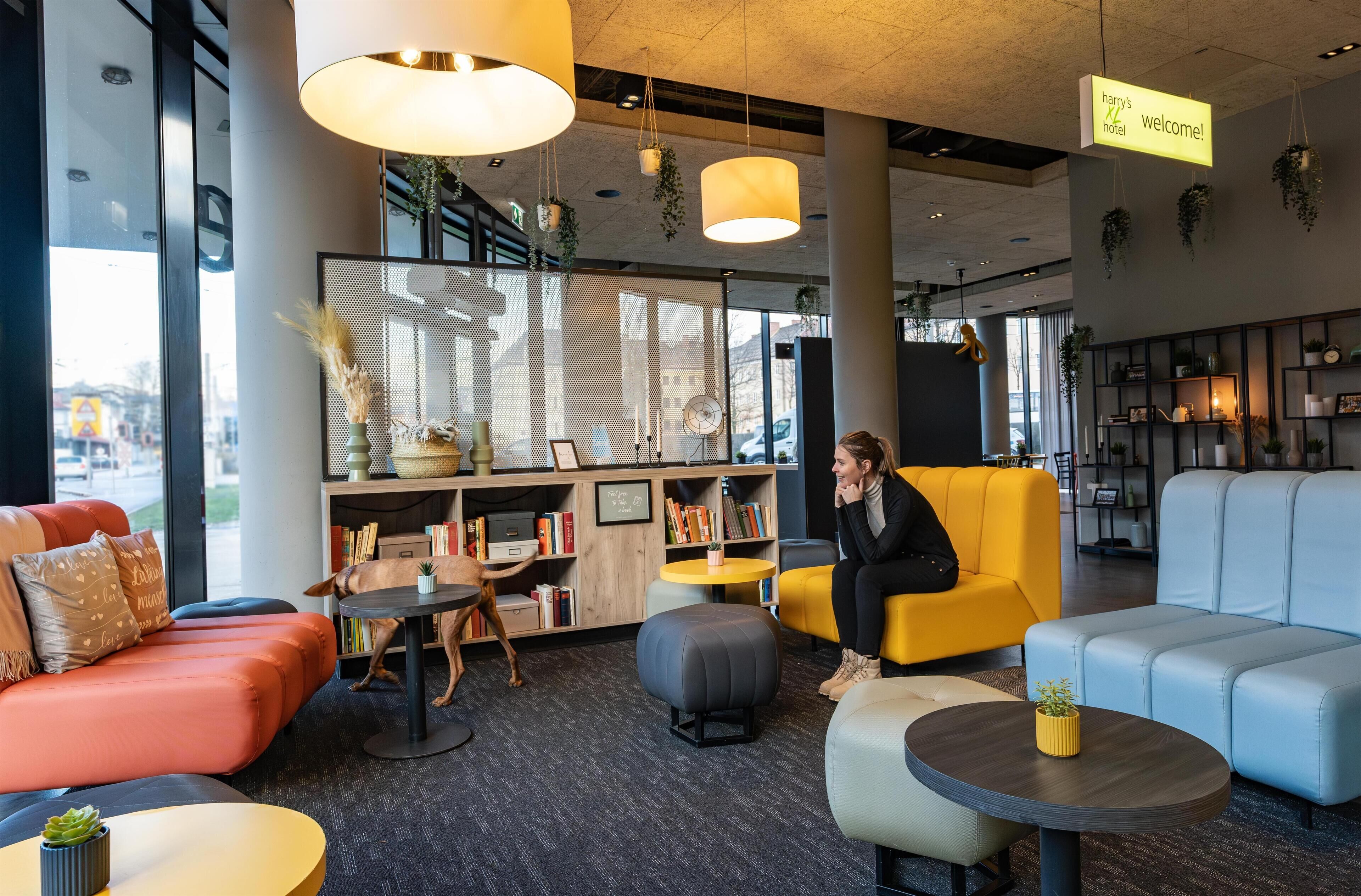 A modern hotel lobby with seating areas, a reception desk, and a bookshelf.