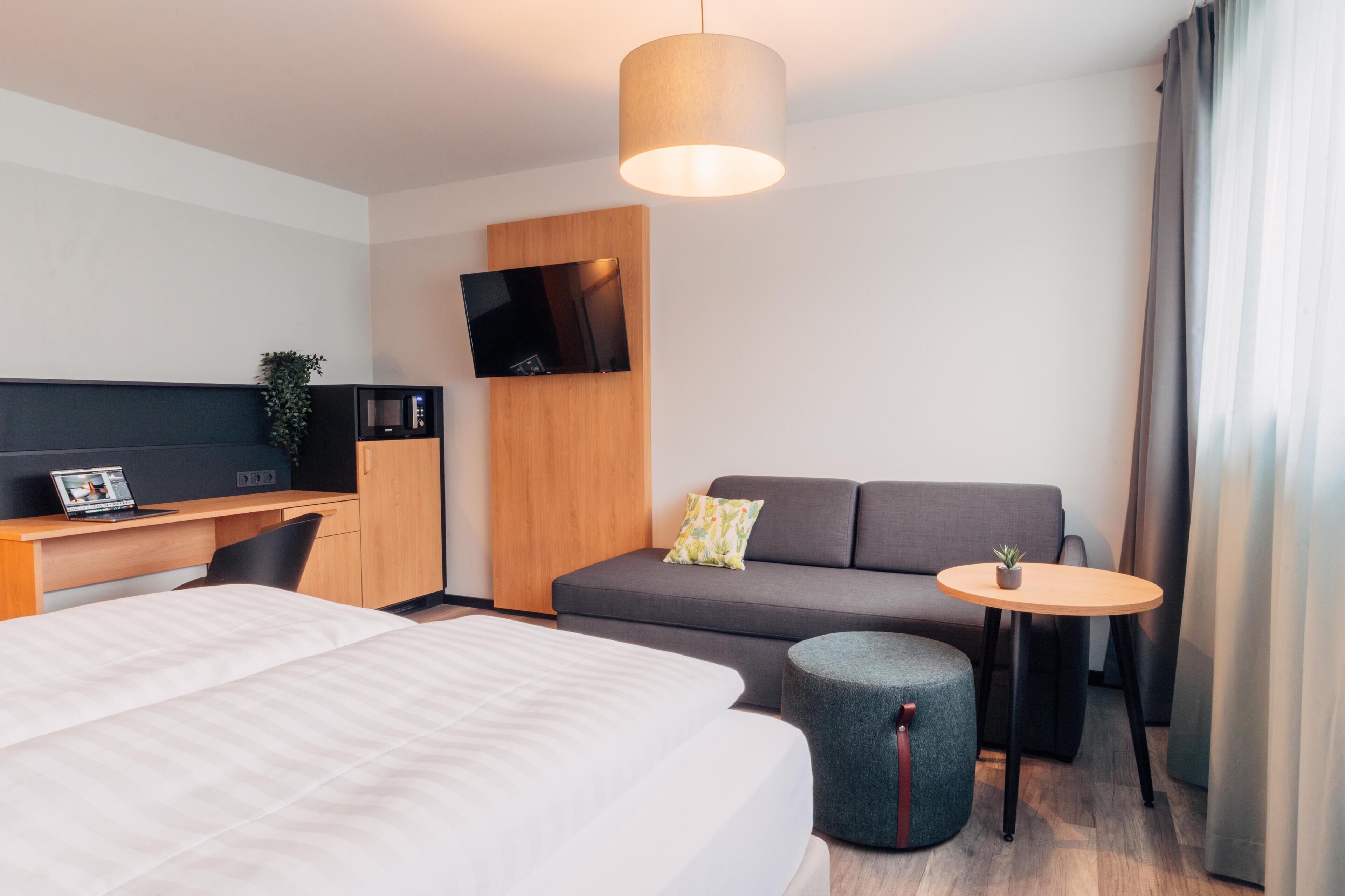 Ein modernes Hotelzimmer mit einem Bett, einem Schreibtisch mit einem Laptop, einer Couch, einem kleinen runden Tisch und einem an der Wand montierten Fernseher.