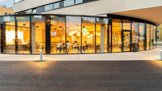 A modern building with a curved glass facade, outdoor seating area, and a well-lit interior. - harry's home Salzburg (Salzburg)