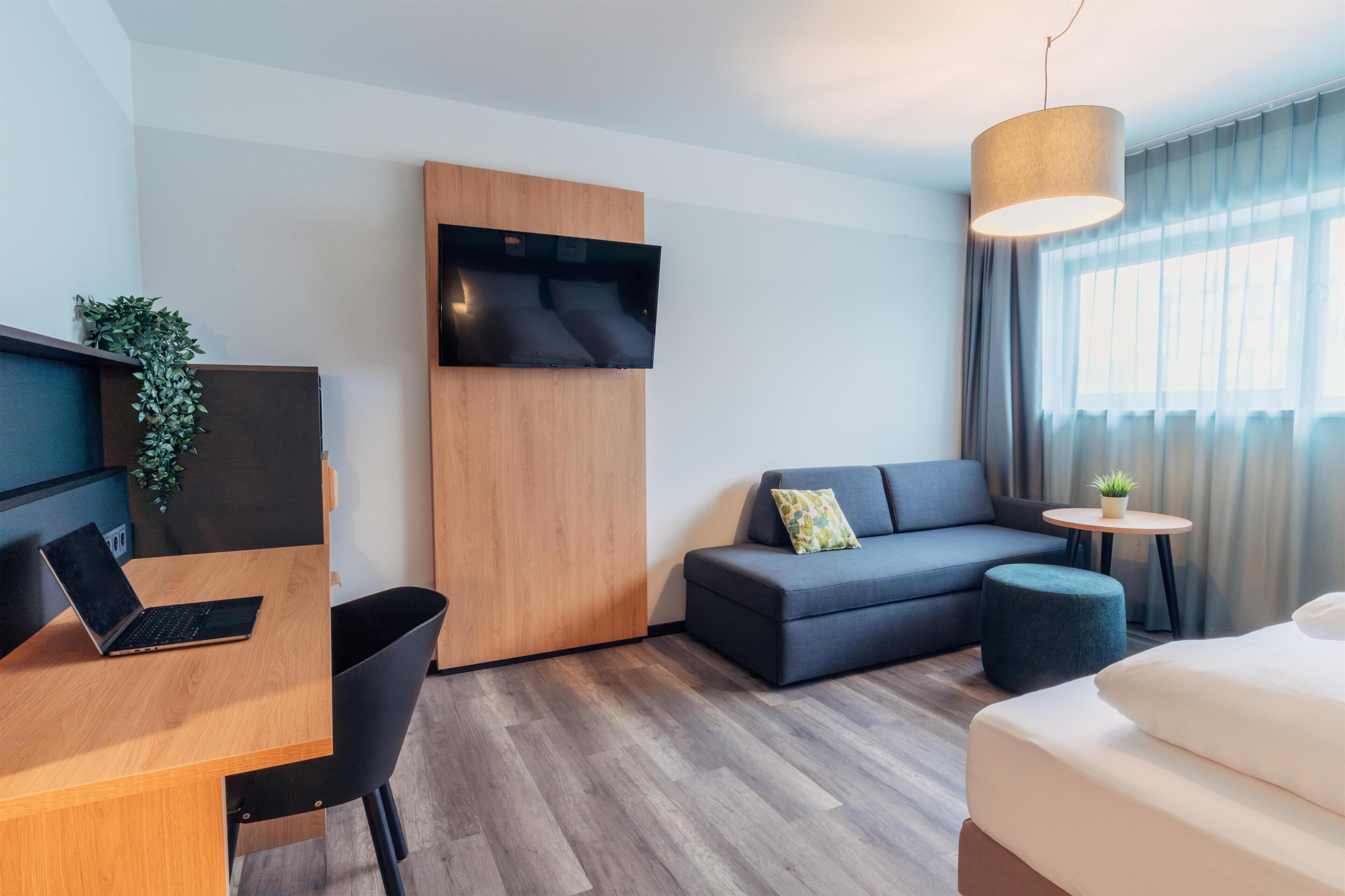 A modern hotel room with a wooden desk, a sofa, a bed, a TV mounted on a wooden panel, and a small round table with a plant.
