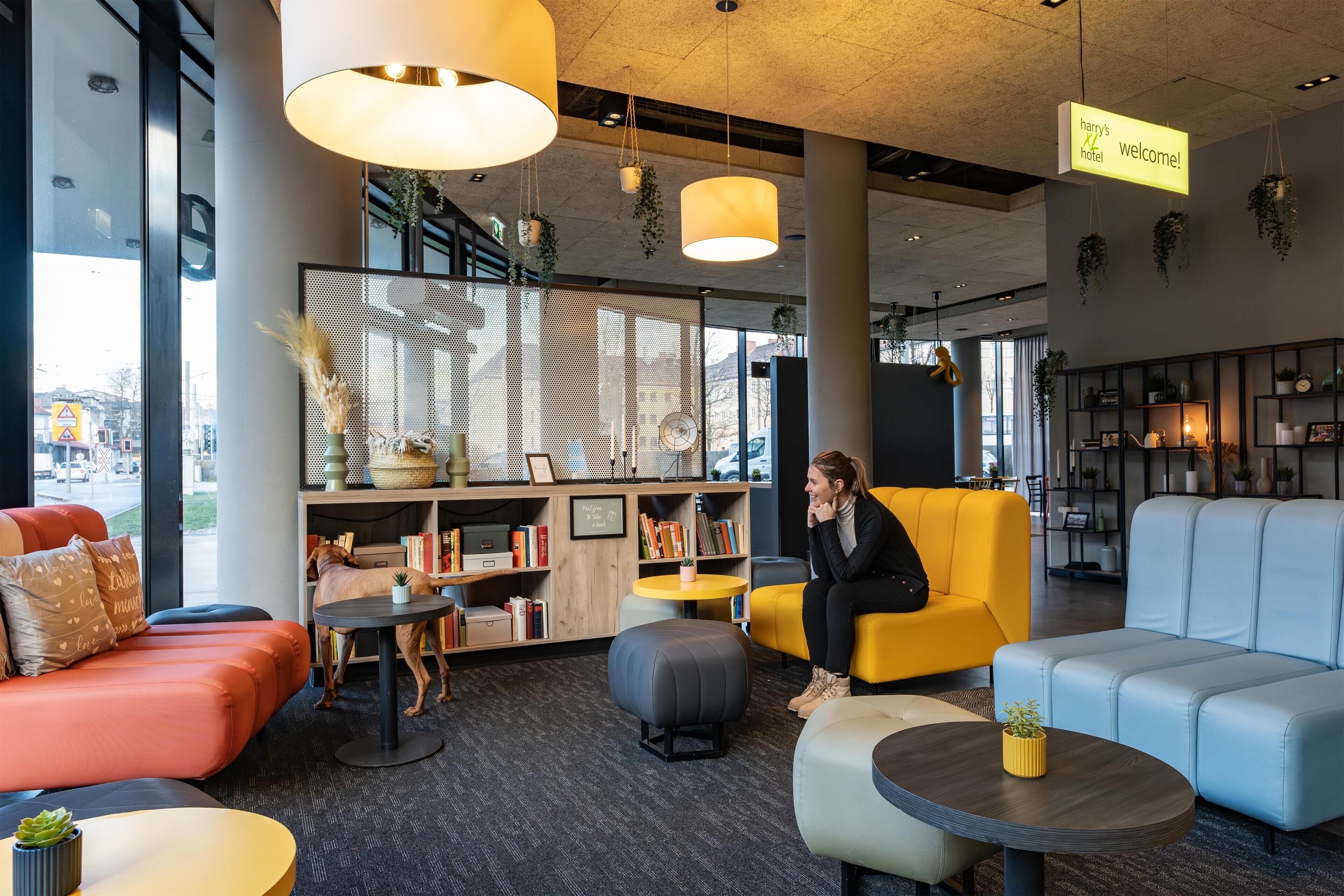 A modern hotel lobby with seating areas, a reception desk, and a large window offering a view of the city.