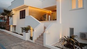 Une maison moderne à deux étages, dotée d’un escalier menant à l’étage supérieur, d’un balcon et d’un éclairage extérieur soigné.