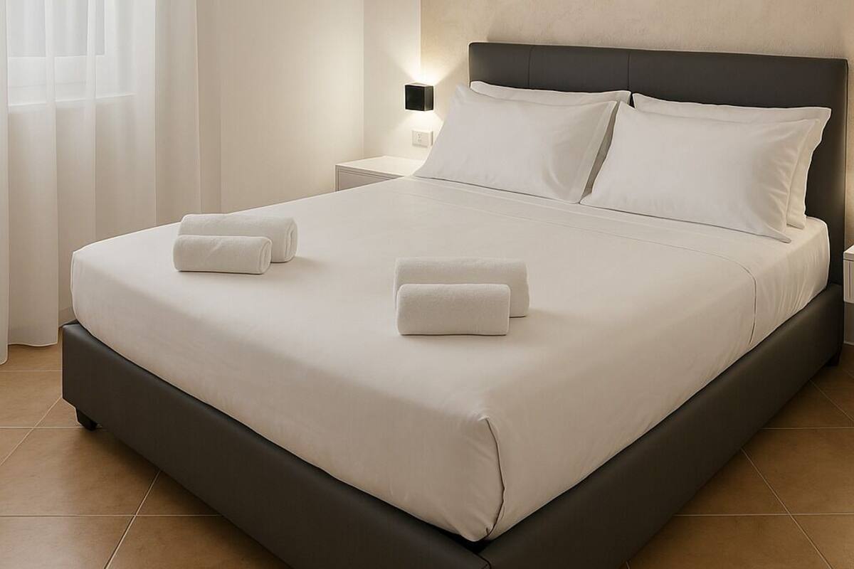 Een net opgemaakt bed met witte lakens en twee handdoeken, een nachtkastje met een lamp en een raam met dunne gordijnen.