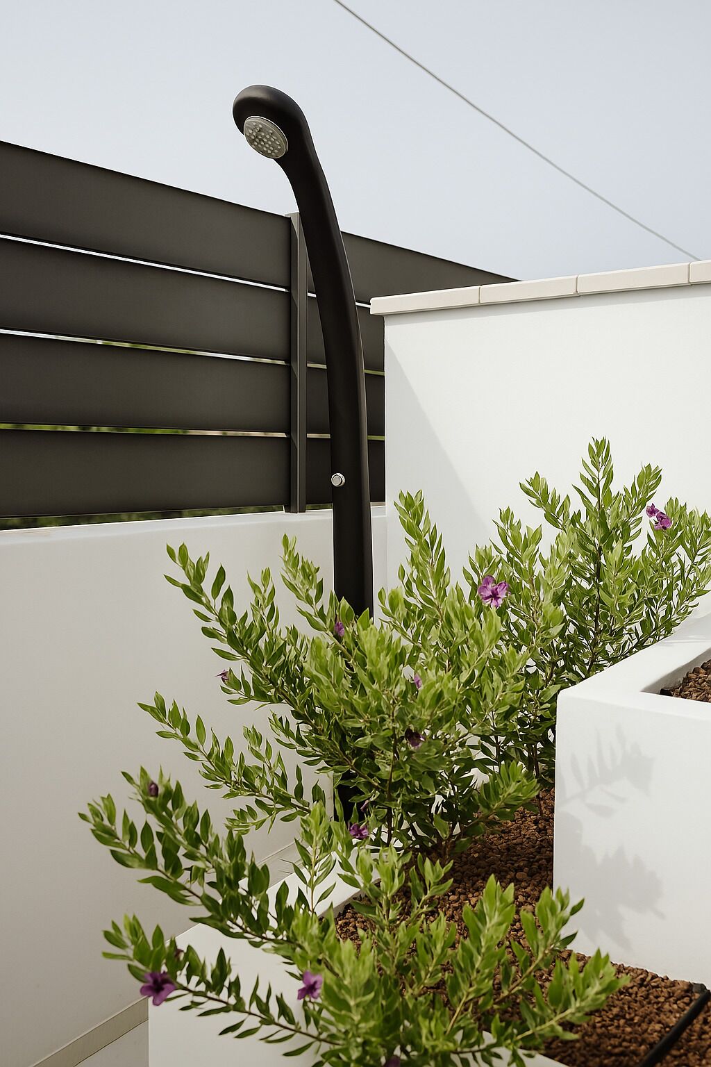 A modern outdoor planter with green foliage and purple flowers, set against a white wall and a dark metal structure.