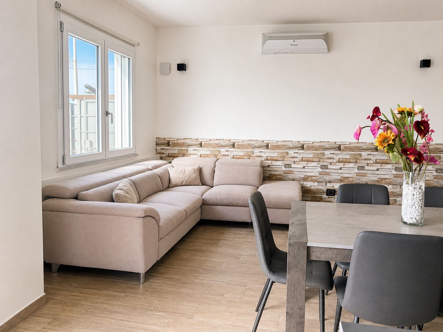 A modern living room with a beige sectional sofa, a dining table with chairs, and a decorative vase with flowers.