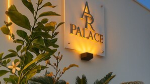 Une enseigne pour « AR PALACE », avec un panneau bien éclairé, entourée d’un jardin agrémenté de citronniers et d’un palmier, et bordée d’un sentier de galets.