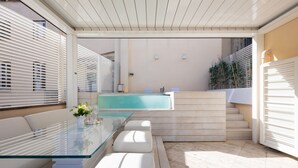 Uma área de estar moderna ao ar livre, com mesa de vidro, bancos brancos acolchoados e piscina ao fundo.