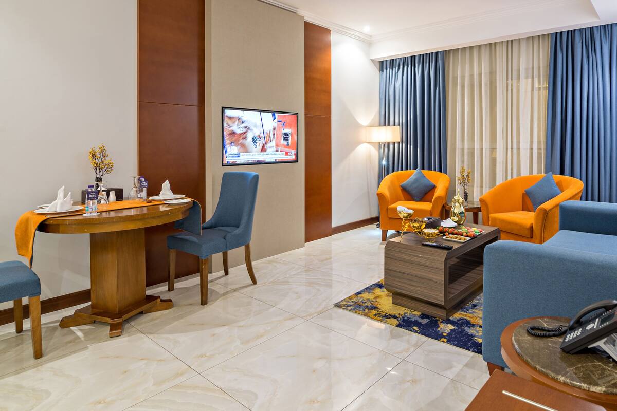 A hotel room with a dining area, two orange armchairs, a blue sofa, a coffee table, and a TV mounted on the wall.