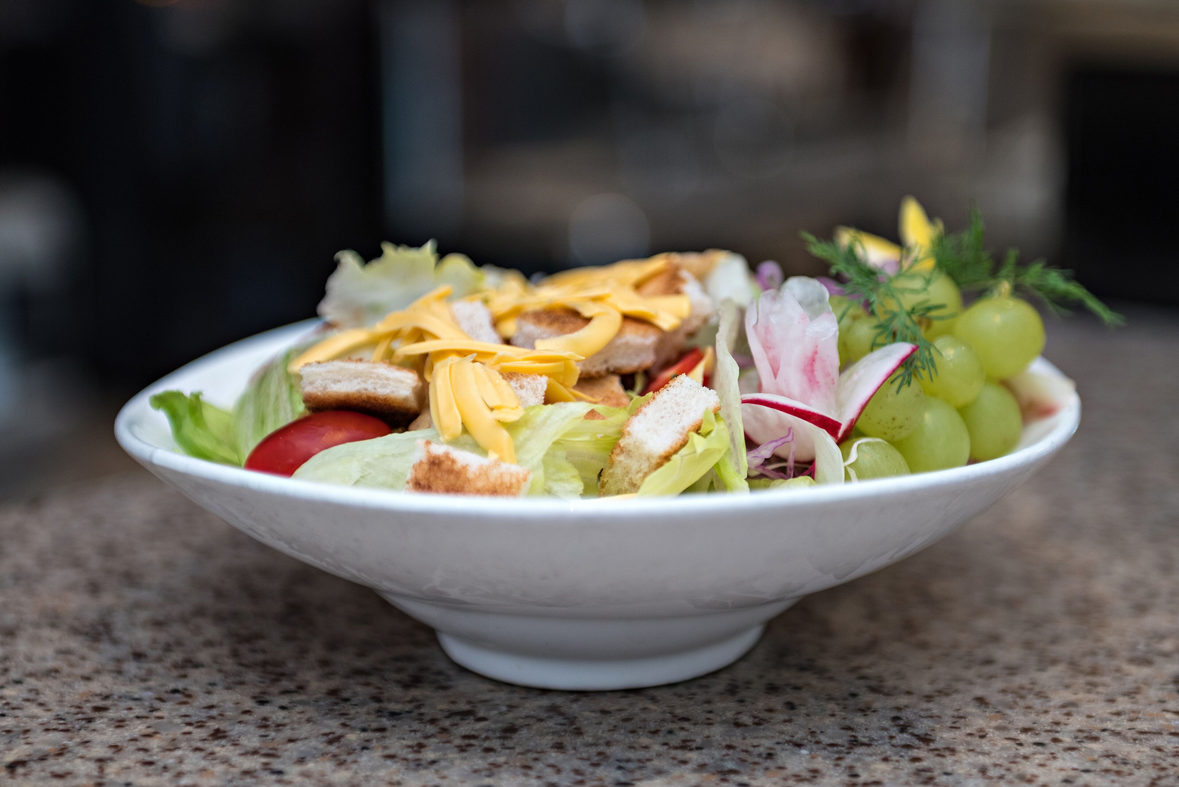 A white bowl containing a salad with lettuce, tomatoes, radishes, and grapes, placed on a speckled countertop.