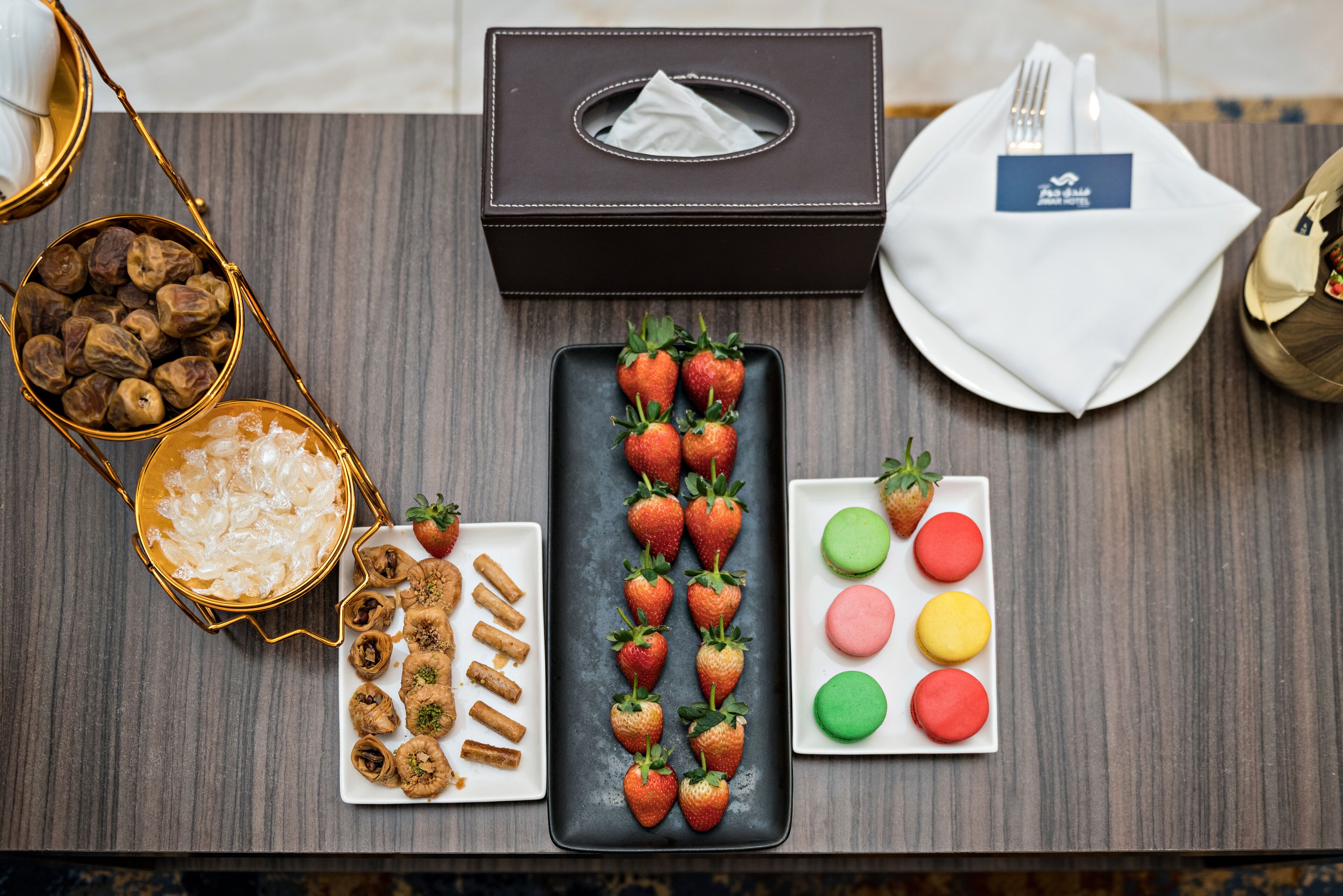 A table with a tissue box, a plate of strawberries, a plate of macarons, a bowl of nuts, and a plate of cookies.