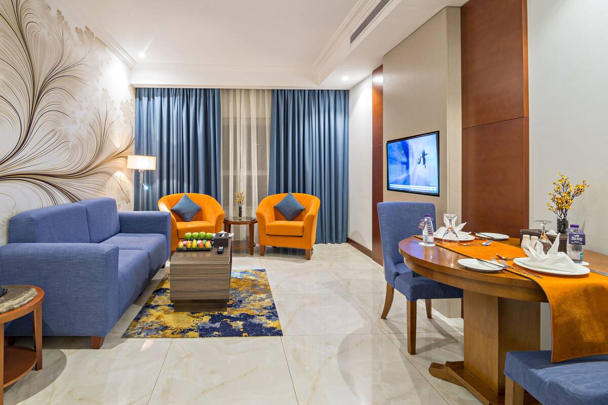 A modern hotel room with a blue sofa, orange armchairs, a dining table set for two, and a large TV on the wall.