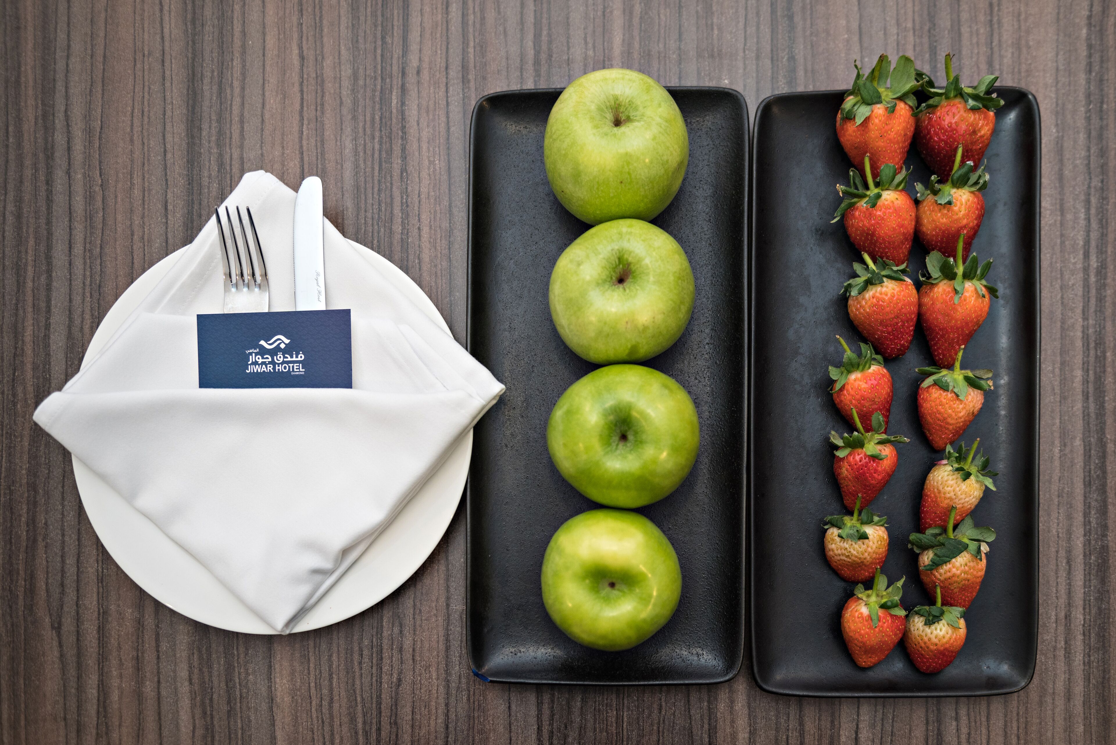 A neatly arranged table with a folded white napkin, a set of cutlery, and two rectangular plates. One plate has three green apples, and the other has a row of strawberries.