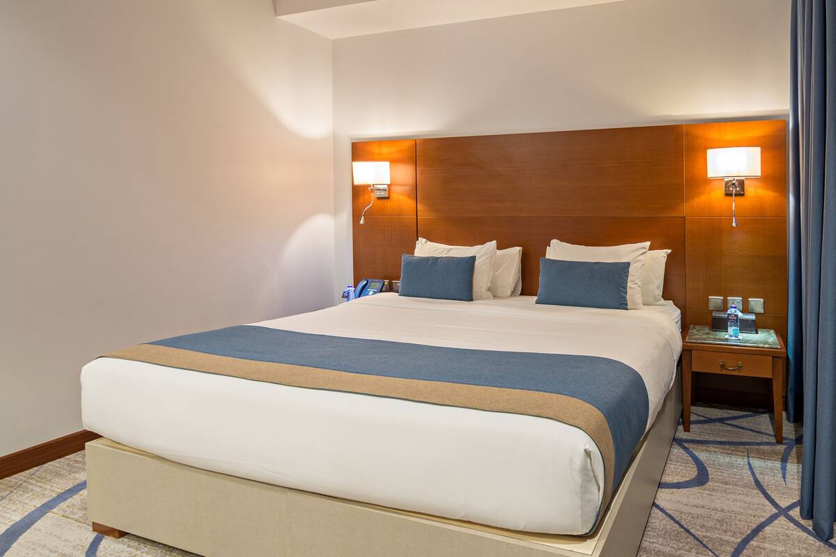 A hotel room with a large bed, two bedside tables, a headboard with integrated lighting, and a patterned carpet.