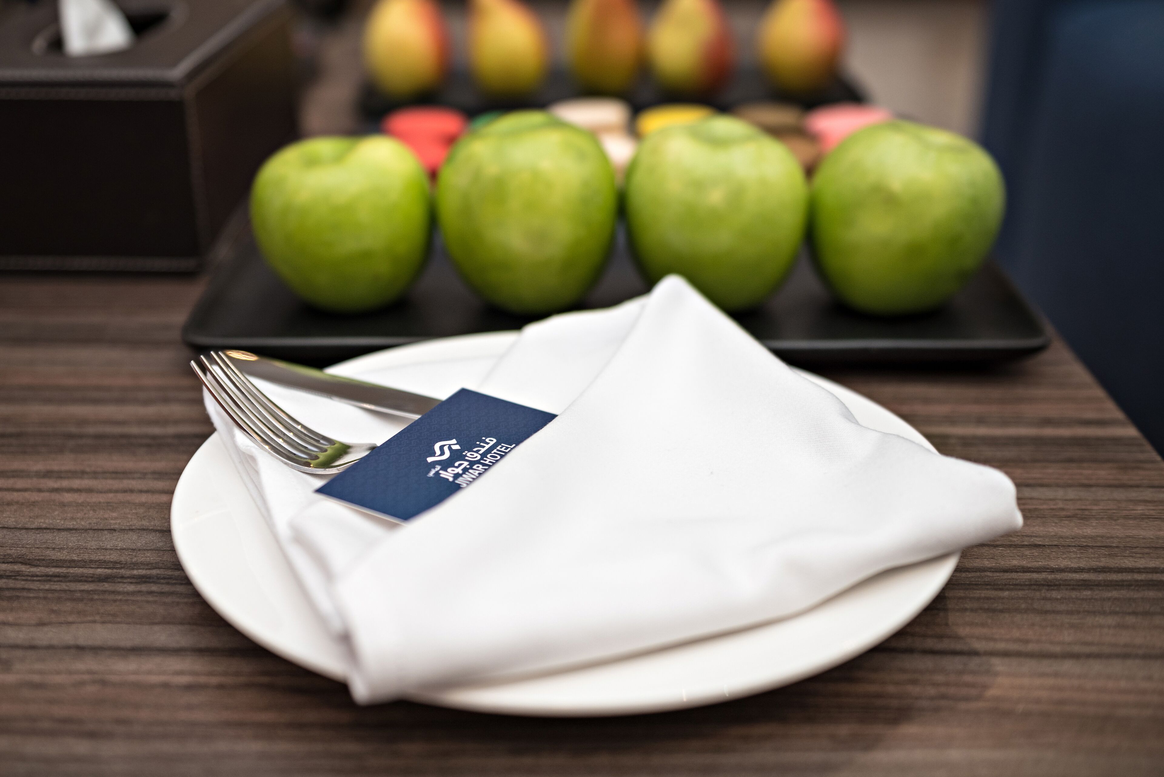 Een netjes gedekte eettafel met een gevouwen servet, een vork en een bord op een houten tafel. Op de achtergrond staan een aantal groene appels op een zwarte schaal.