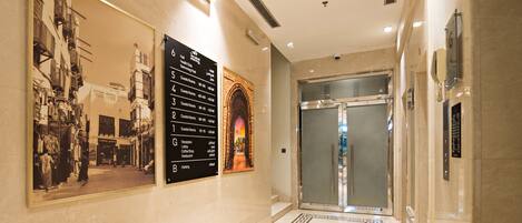 Corredor de hotel com piso de mármore, balcão de recepção e elevador de vidro.