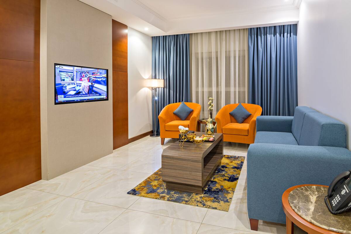A modern hotel room with a blue sofa, orange armchairs, a coffee table with decorative items, a flat-screen TV on the wall, and a telephone on a side table.