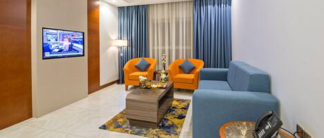 A modern hotel room with a blue sofa, orange armchairs, a coffee table with decorative items, a flat-screen TV on the wall, and a telephone on a side table.