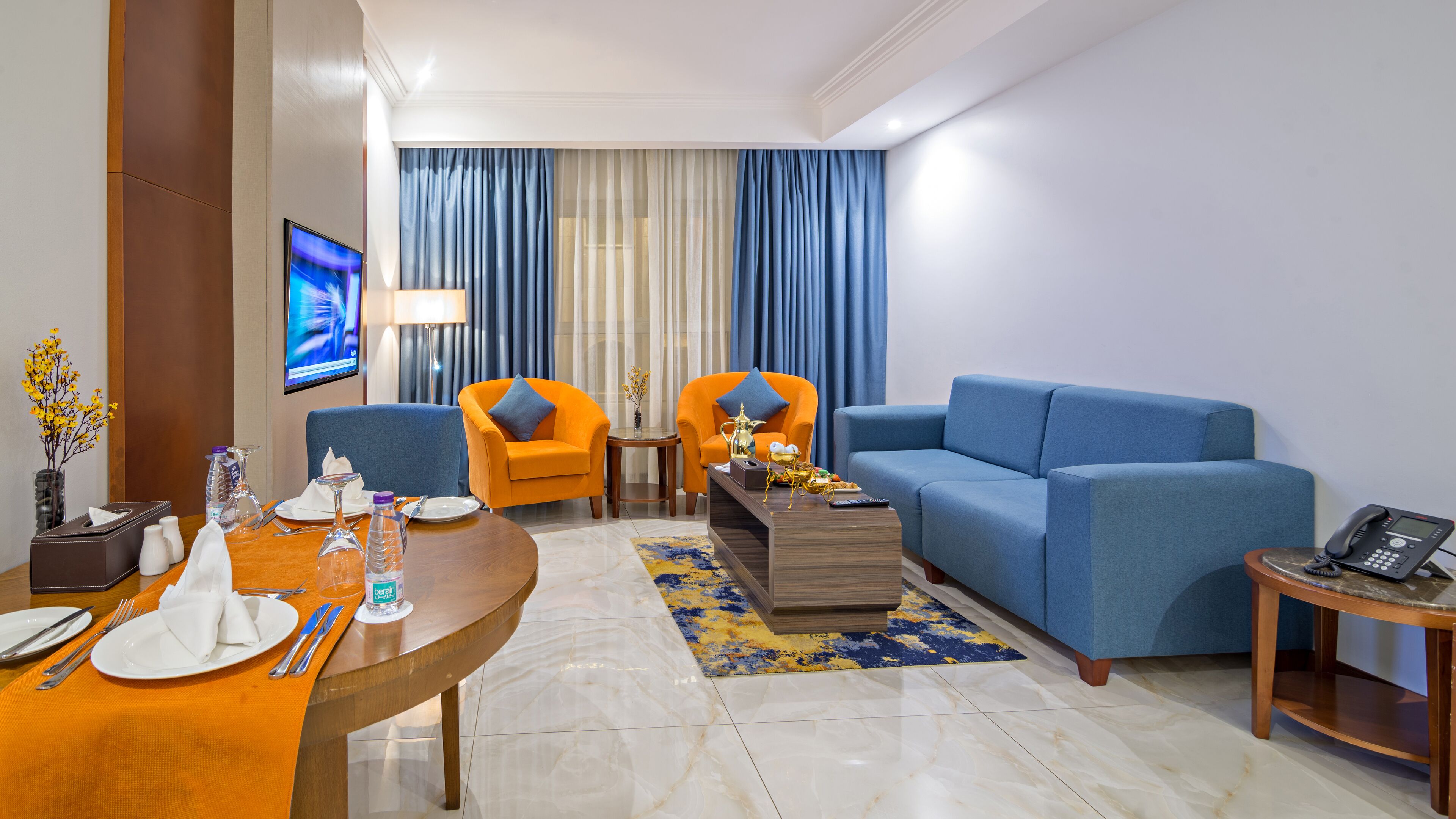 A modern living room with a blue sofa, orange armchairs, a wooden coffee table, and a small side table with a phone.