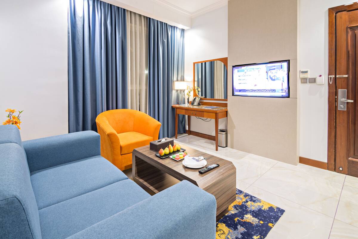 A modern hotel room with a blue sofa, an orange armchair, a wooden coffee table with fruit and a remote control, a desk with a lamp, a TV, and a wooden door.