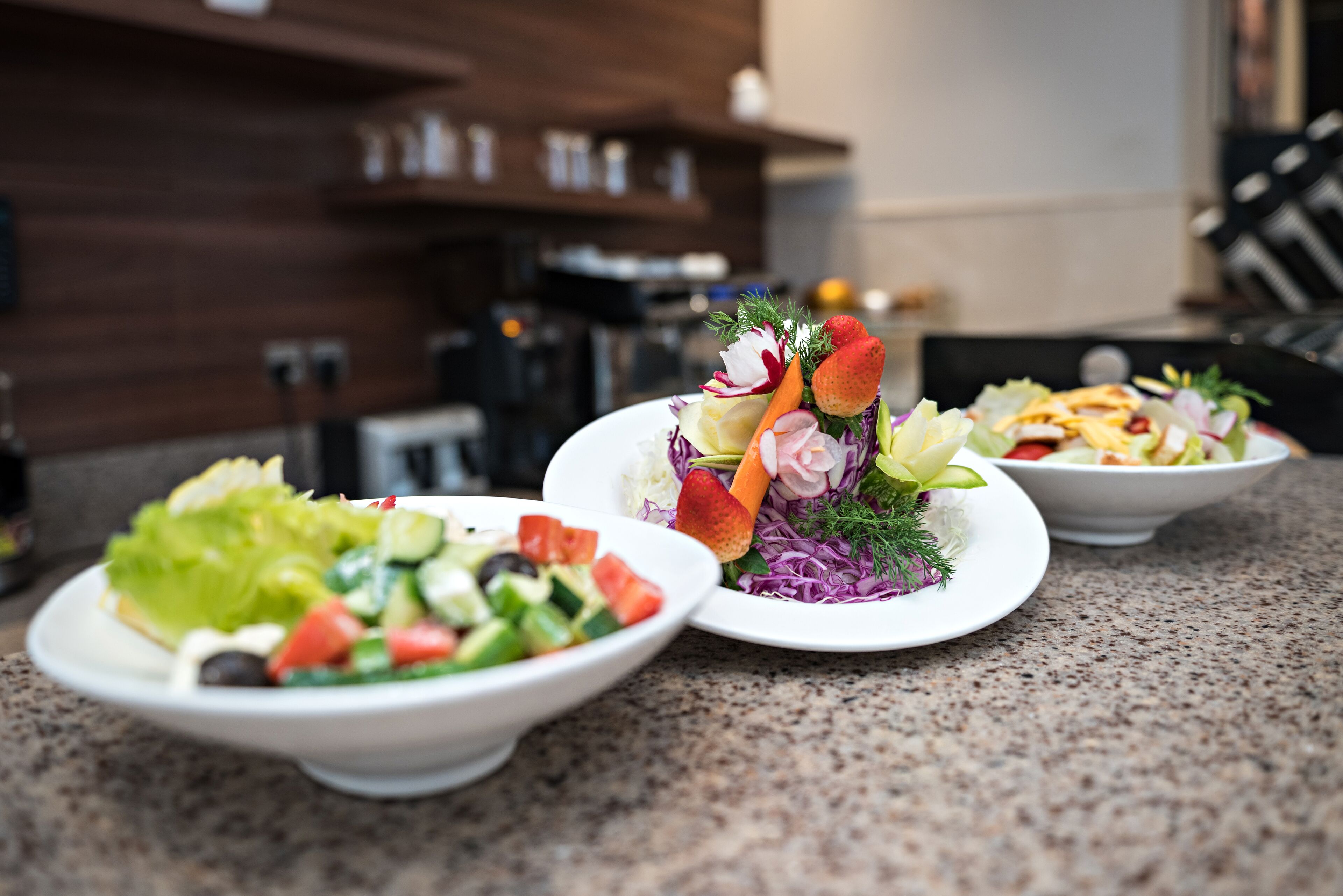 Three bowls of salad on a granite countertop with a kitchen in the background.