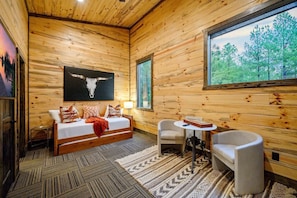 Quarto de cabana de toras com cama, duas cadeiras, uma mesa e uma janela grande.