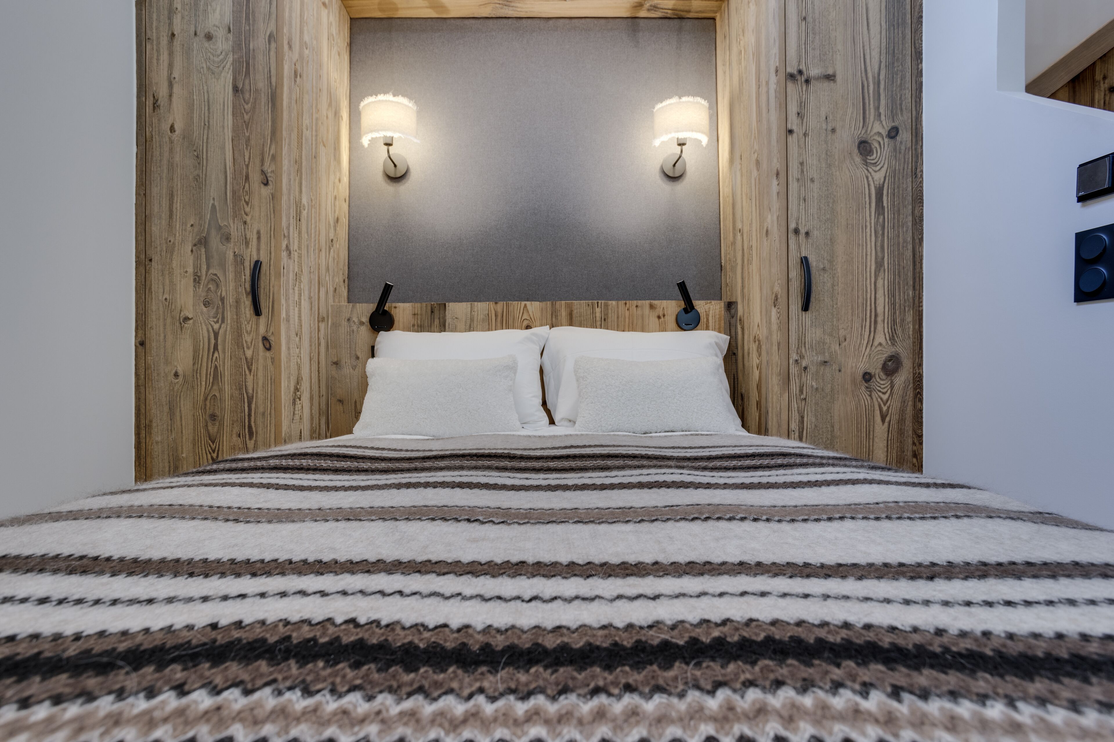 A bed with a striped blanket, wooden headboard, and two wall-mounted lamps.