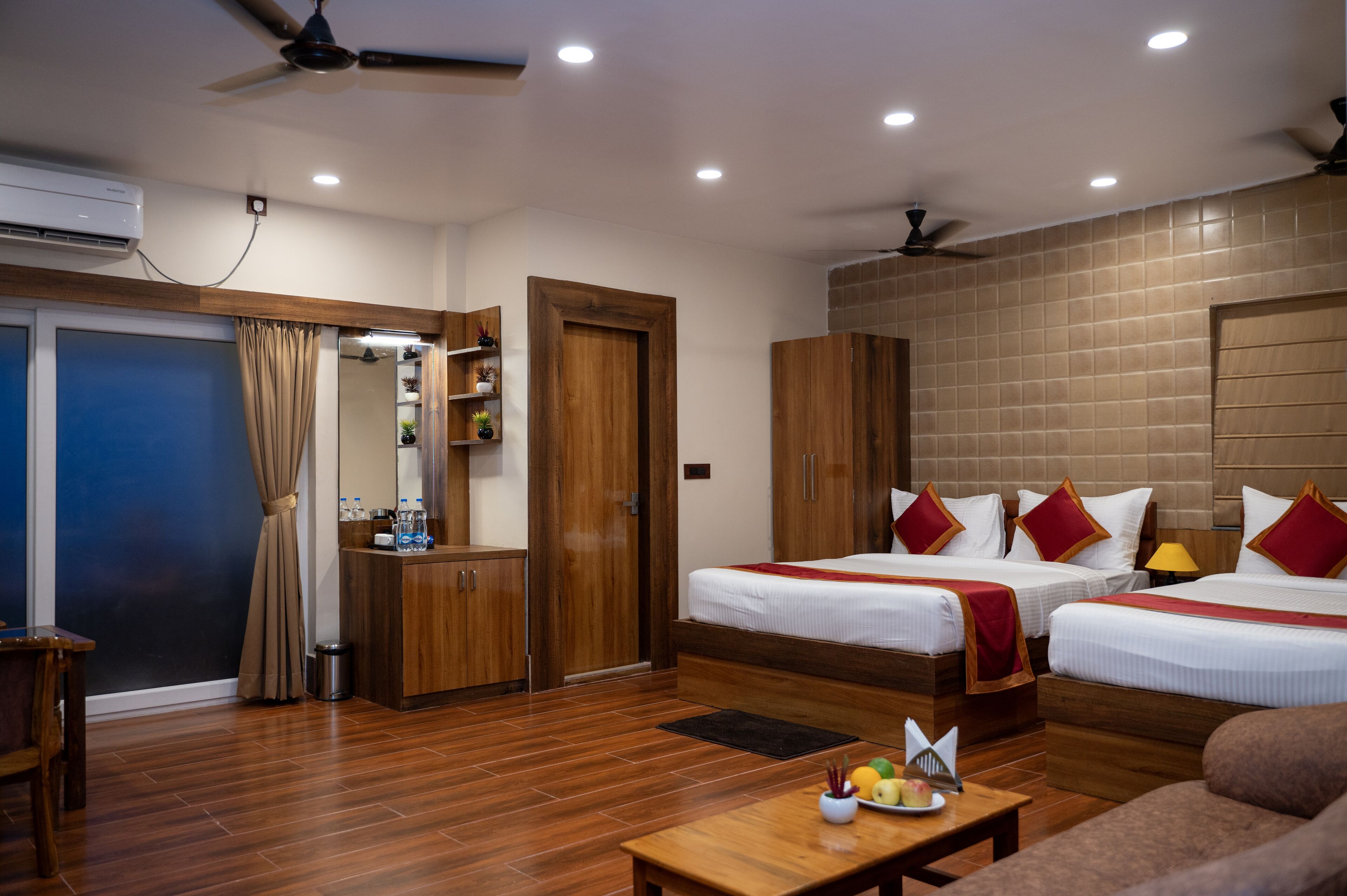 A hotel room with two beds, wooden furniture, a desk, and a small table with fruits and a bottle.
