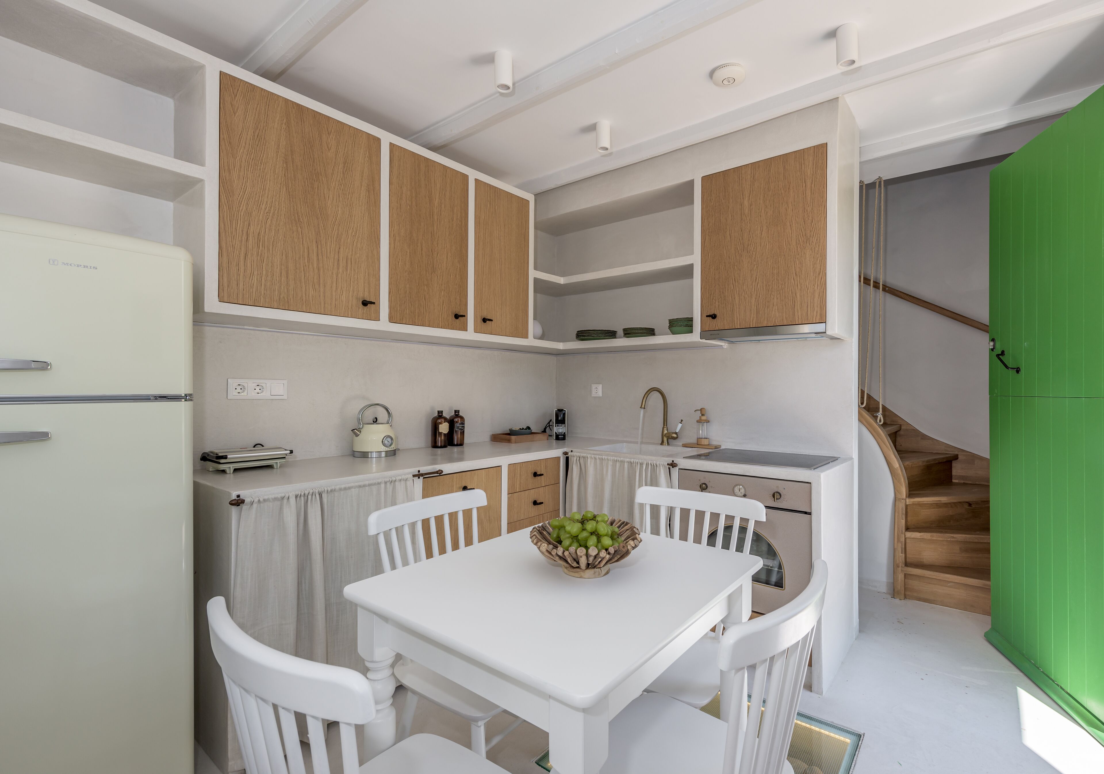 Cocina compacta con muebles blancos, una mesa de comedor con sillas, un frutero y una puerta verde.