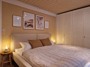 A bedroom with a large bed, a bedside table with a lamp, a wardrobe, and three framed pictures on the wall.
