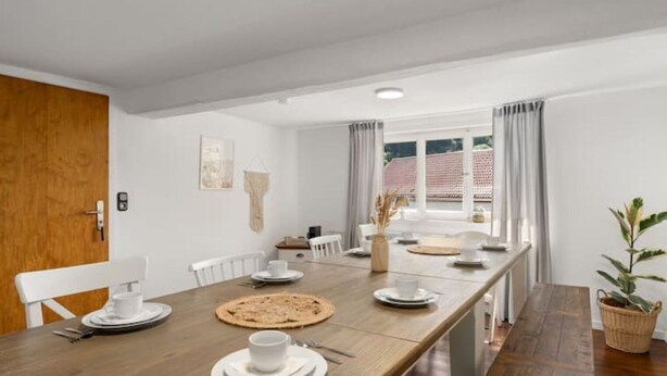A modern dining area with a long wooden table, white chairs, and a window with curtains. - Cozy vacation home Zur Alten Burg/Family reunion/Edersee (Vöhl)