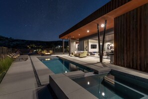 A modern outdoor pool area with a clear night sky, a covered seating area, and a well-lit interior space. - Canyon Ridge by Fieldtrip | Modern Indoor/Outdoor Oasis w Views (Yucca Valley)