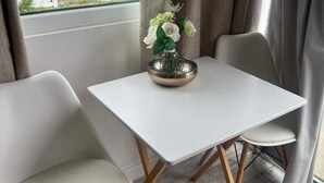A small, modern room with a white table and two chairs, a vase with flowers, and a view of a white fence outside. - Stunning One Bed Modern & Contemporary Chalet (Bridlington)