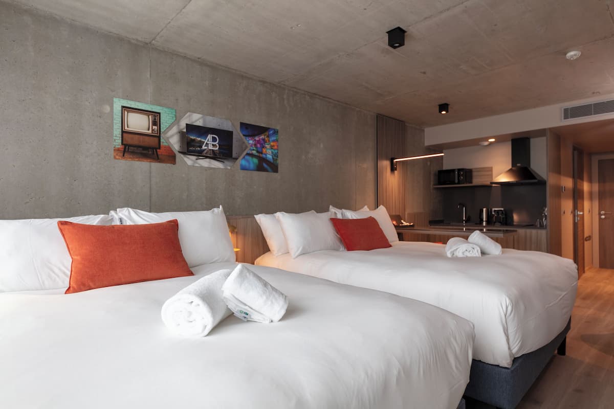 A hotel room with two beds, white bedding, red pillows, a kitchen area, and artwork on the wall.