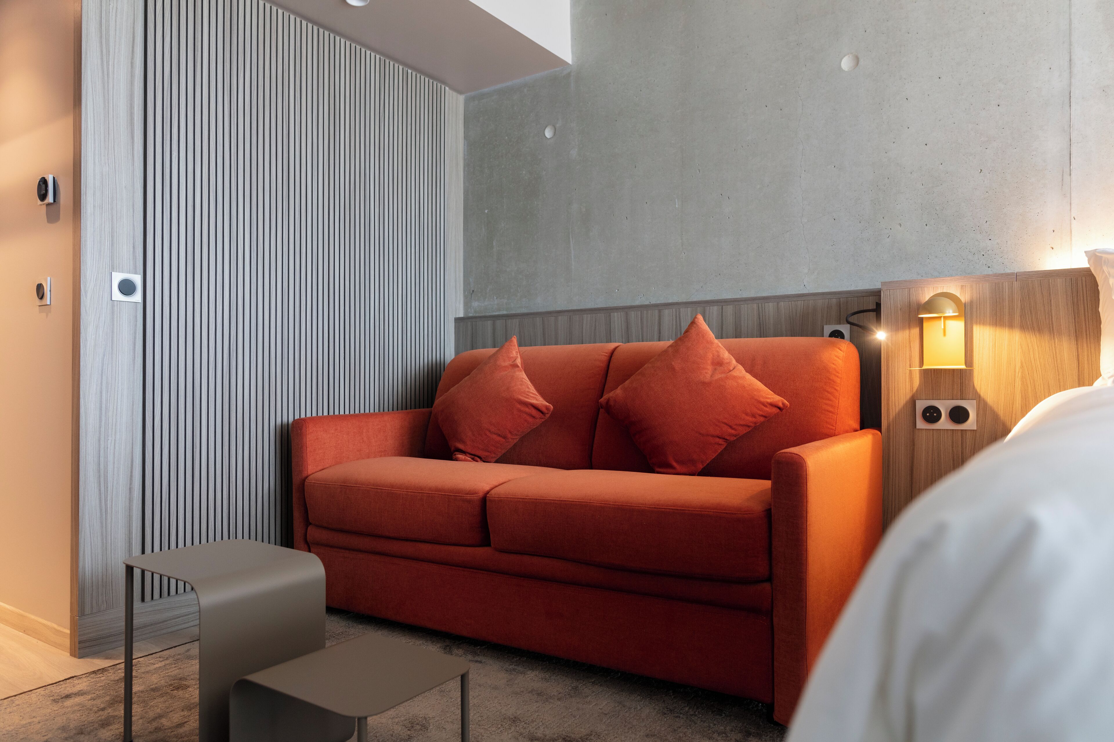 A modern hotel room with a red sofa, a small side table, and a wall-mounted light fixture.