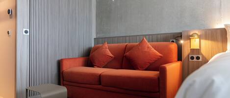 A modern hotel room with a red sofa, a small side table, and a wall-mounted light fixture.