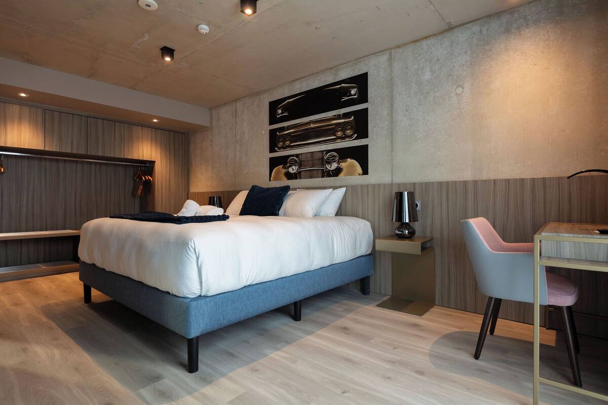 A modern bedroom with a large bed, a desk, and a chair. There is a wall-mounted shelf with decorative items.