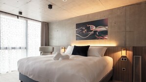 Una habitación de hotel moderna con una cama grande, un cabecero con una lámpara y un cuadro colgado en la pared, además de una ventana con cortinas.