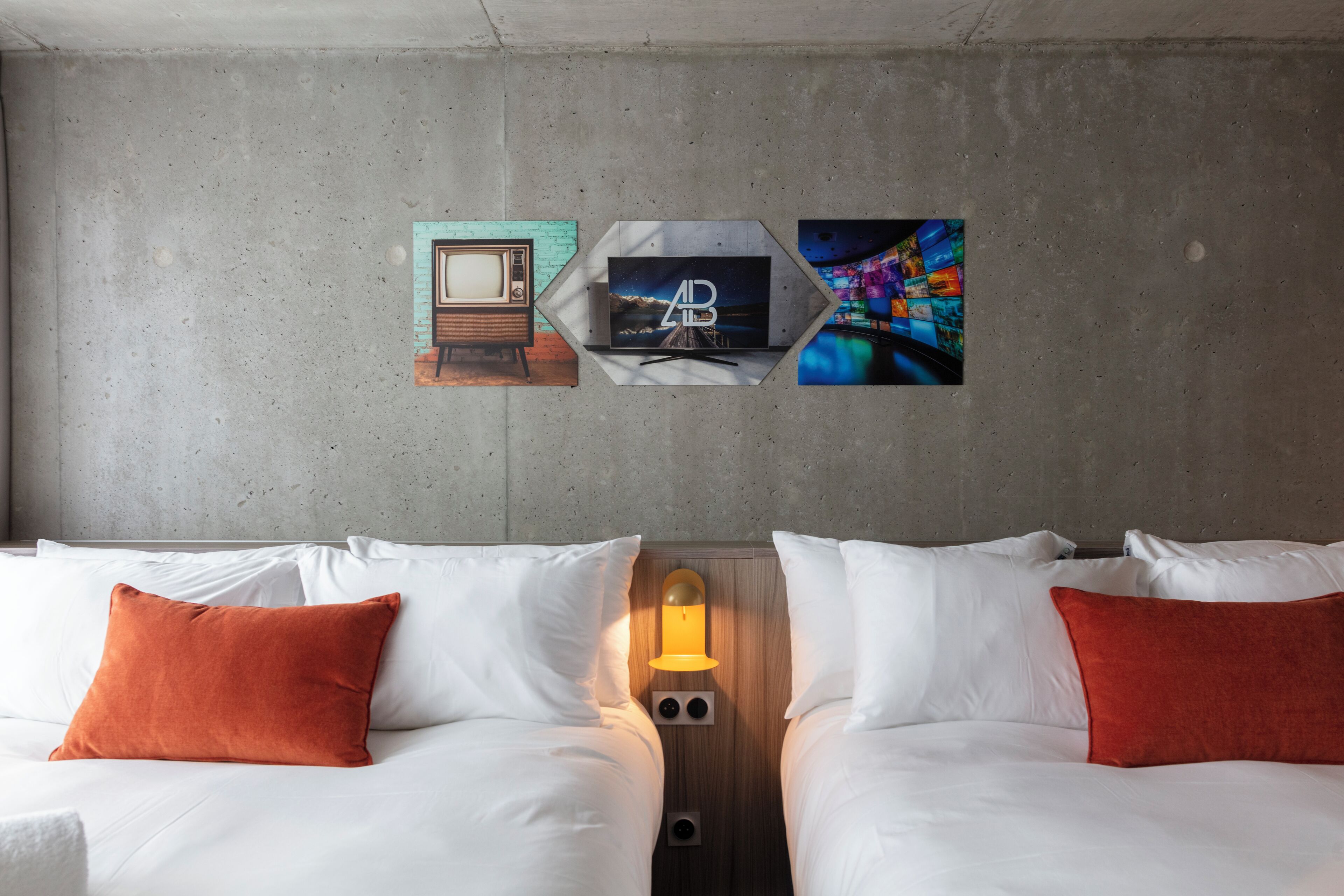 A hotel room with two beds, white bedding, and orange pillows. There are wall-mounted artworks depicting a vintage TV, a mountain landscape, and a colorful abstract design.