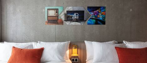 A hotel room with two beds, white bedding, and orange pillows. There are wall-mounted artworks depicting a vintage TV, a mountain landscape, and a colorful abstract design.