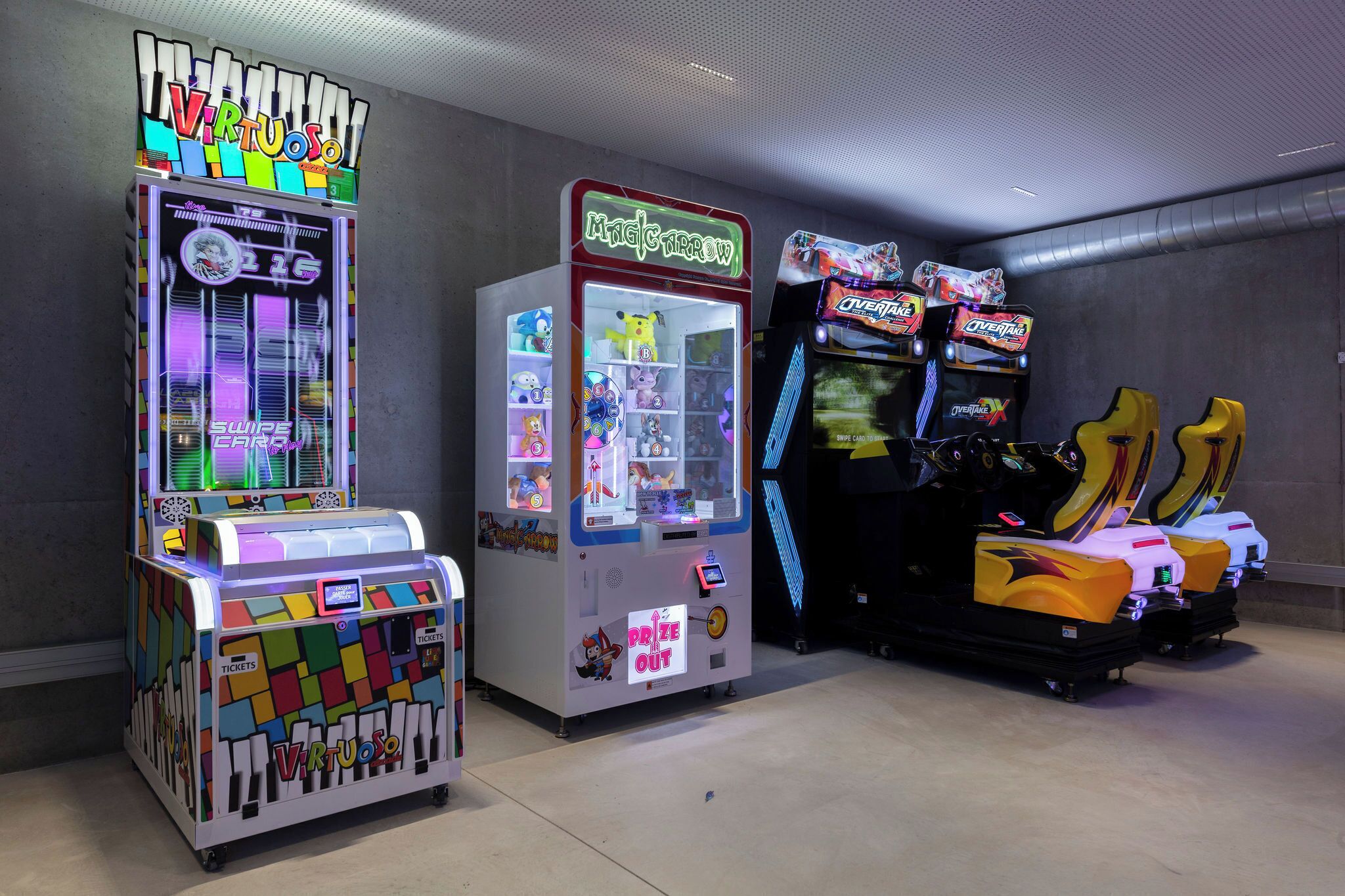Una hilera de juegos de arcade, incluyendo 'Virtuosos', 'Magic Throw' y 'Overtake', en una sala con suelo de concreto y paredes grises.