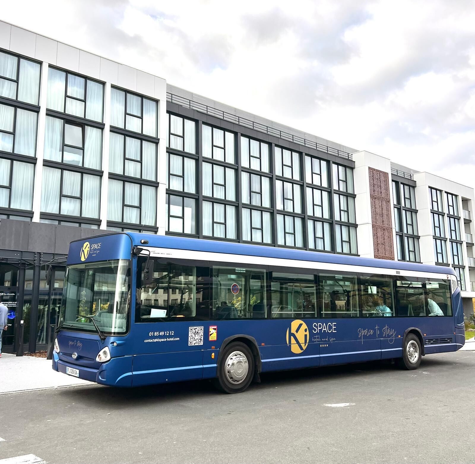 Un autobús azul con el logo 'SPACE' y el lema 'space to stay' estacionado frente a un edificio moderno con ventanas grandes y una columna de ladrillo.