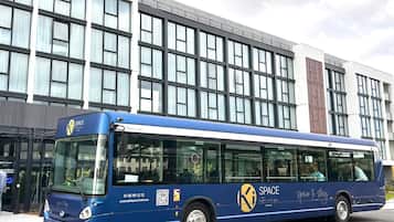 Un autobús azul con el logo 'SPACE' y el lema 'space to stay' estacionado frente a un edificio moderno con ventanas grandes y una columna de ladrillo.