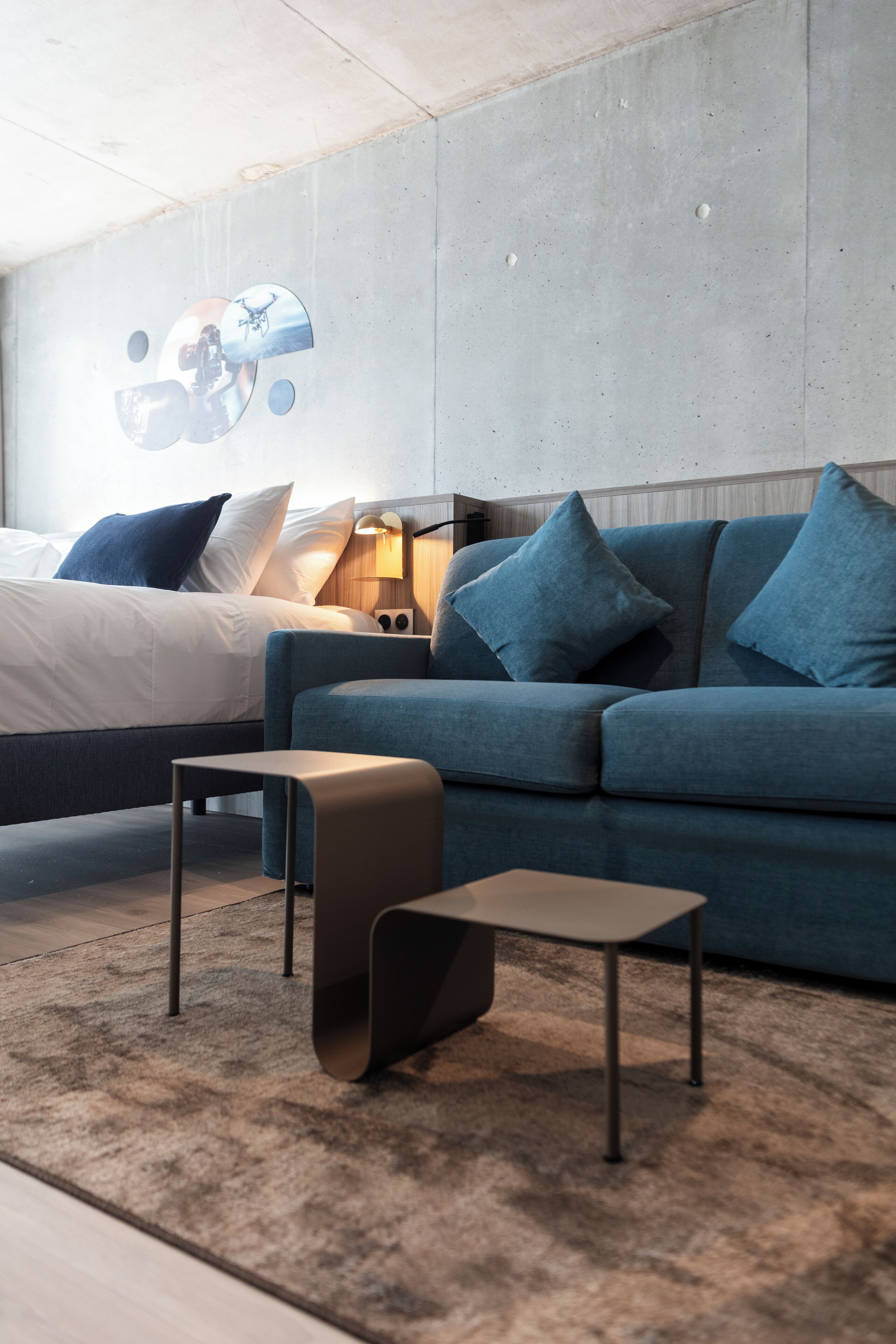 A modern hotel room with a bed, a blue sofa, a small table, and a wall-mounted artwork.