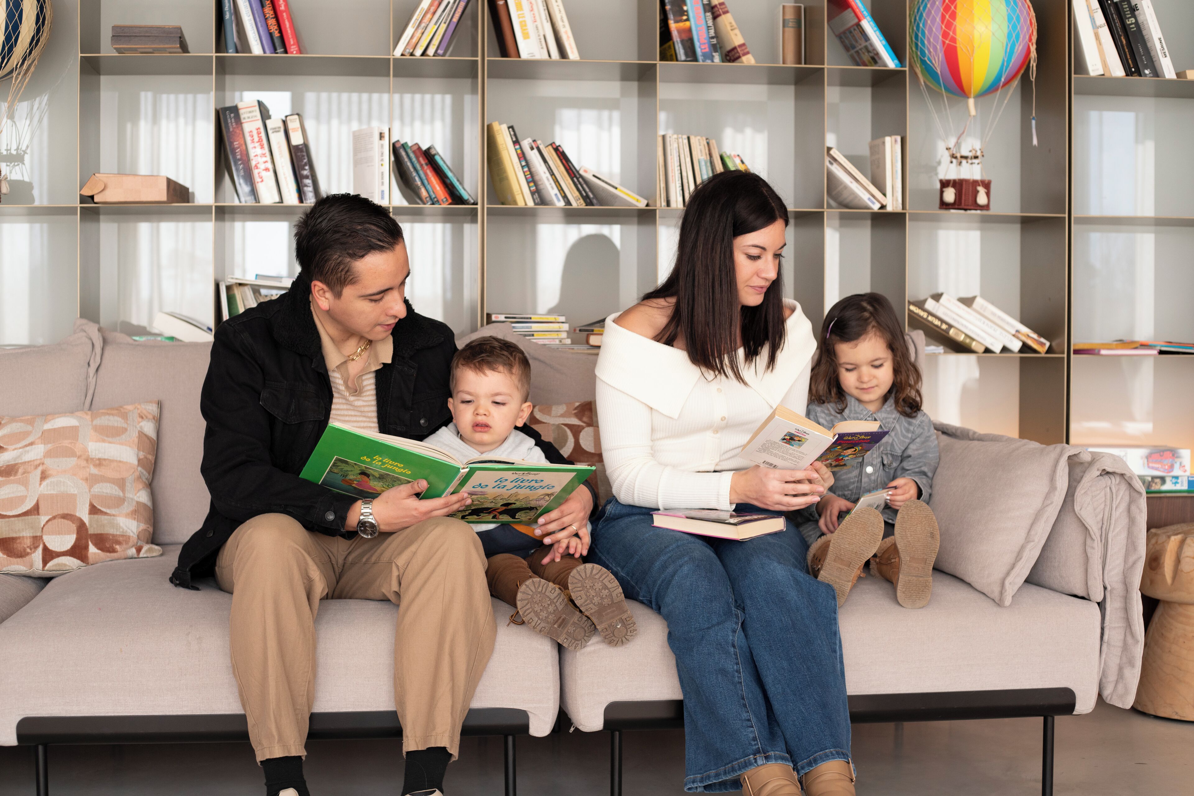 Eine vierköpfige Familie sitzt auf einem Sofa mit Büchern, im Hintergrund steht ein Bücherregal und eine Heißluftballon-Dekoration ist zu sehen.