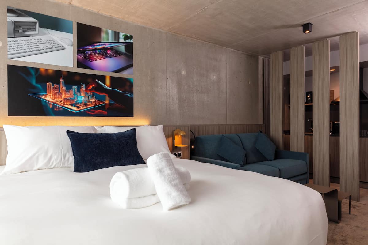 A hotel room with a large bed, a desk with a computer, a blue sofa, and a cityscape artwork on the wall.