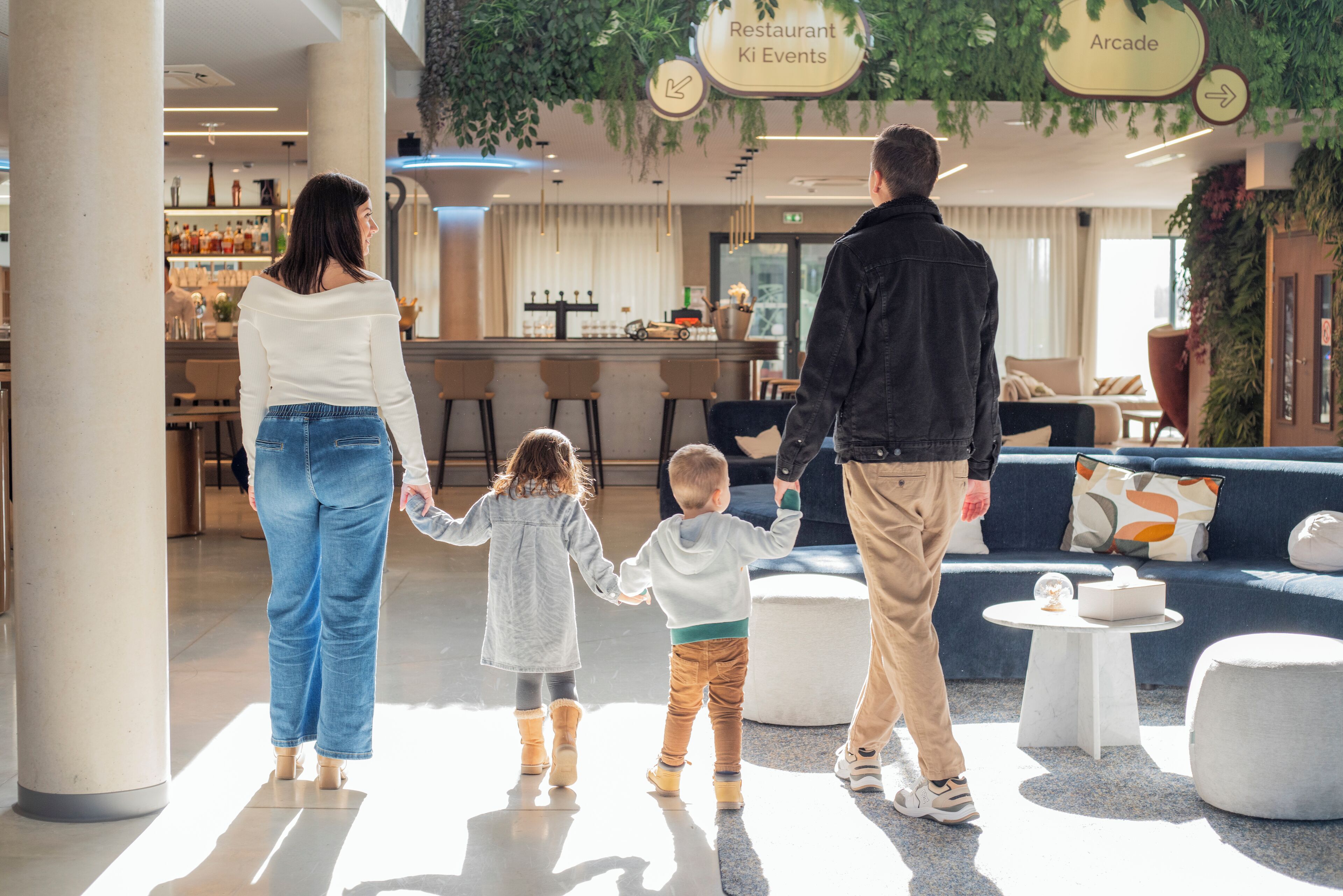 Une famille se promène dans un hall d’entrée moderne, qui comprend un bar, des espaces de détente et des panneaux indicateurs pour accéder au restaurant et à la salle d’arcade.