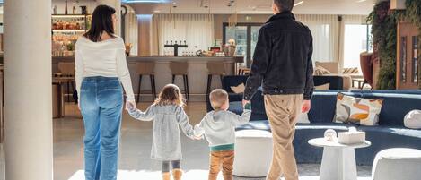 Uma família caminhando por um hall moderno, com área de bar, assentos e placas indicativas para o restaurante e o fliperama.