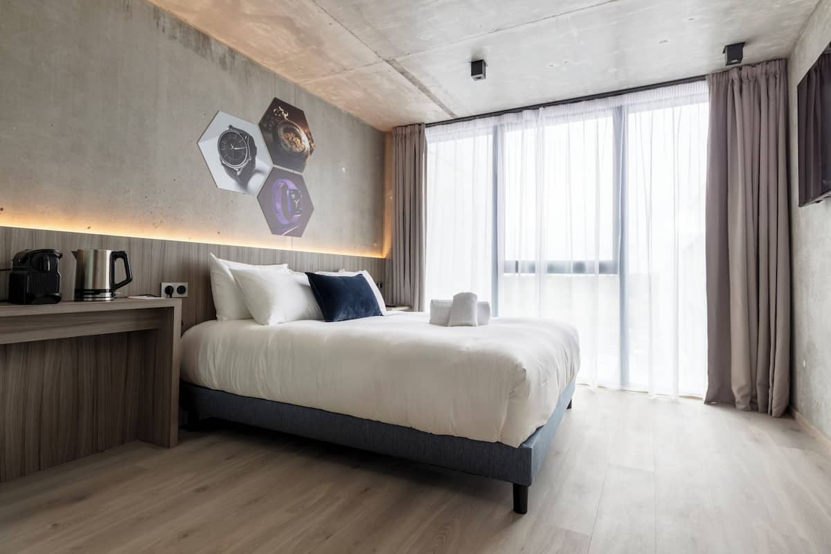 A modern hotel room with a large bed, a wooden bedside table with a coffee maker, and a wall-mounted clock.