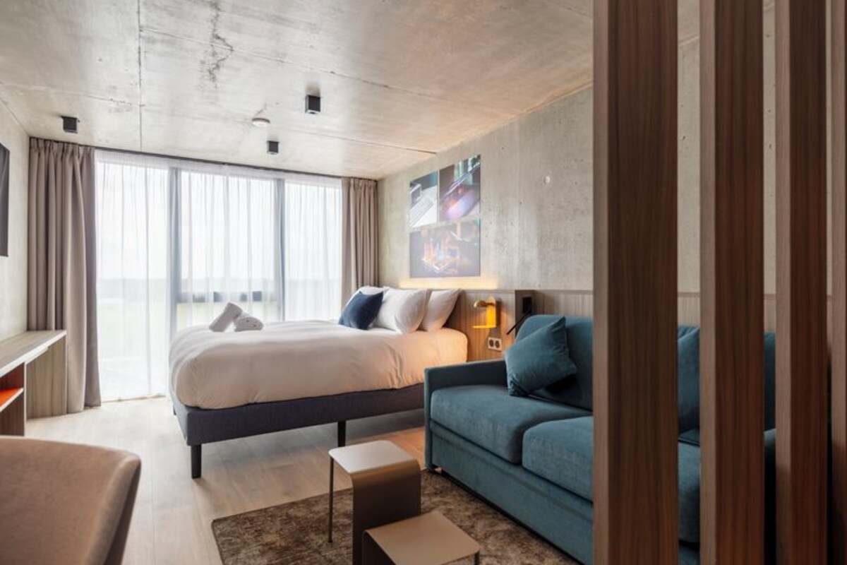 A modern hotel room with a large bed, a sofa, a small table, and a large window.